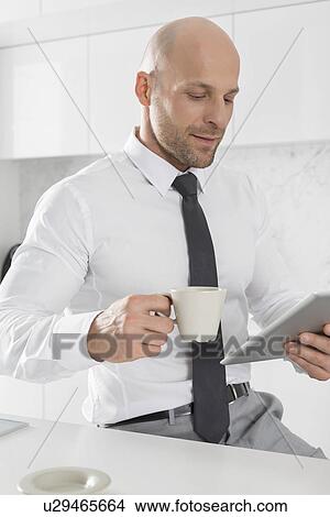 Picture - Mid adult businessman having coffee while using tablet PC in kitchen. Fotosearch