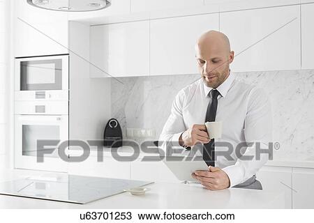 Mid adult businessman having coffee while using tablet PC at kitchen counter View Large Photo Image Stock Image - Mid adult businessman having coffee while using tablet PC at kitchen counter. Fotosearch