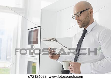 Stock Image - Mid adult businessman having coffee while reading newspaper at home. Fotosearch