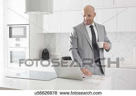 Mid adult businessman having coffee while using laptop in kitchen View Large Photo Image Stock Photo - Mid adult businessman having coffee while using laptop in kitchen. Fotosearch