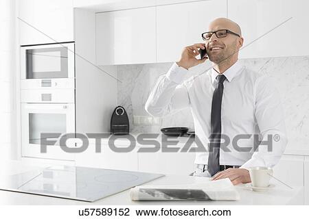 Stock Image - Mid adult businessman on call at kitchen counter. Fotosearch