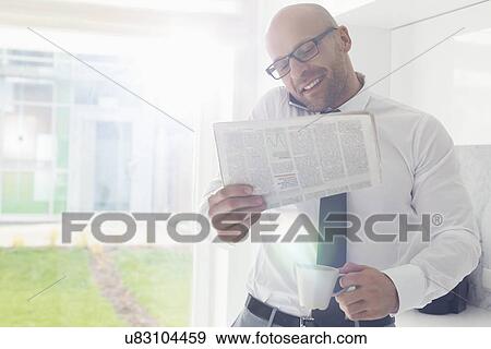 Stock Photo - Mid adult businessman on call while holding newspaper and coffee cup at home. Fotosearch