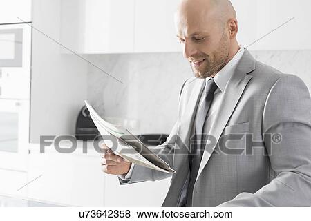 Stock Photo - Mid adult businessman reading newspaper in kitchen. Fotosearch