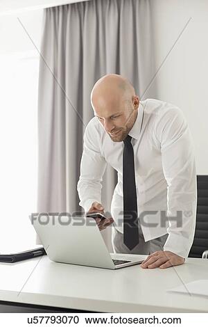 Stock Image - Mid adult businessman using call phone and laptop at table in home office. Fotosearch