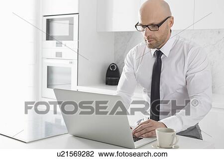 Stock Image - Mid adult businessman using laptop at kitchen counter. Fotosearch