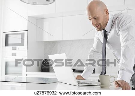 Stock Image - Mid adult businessman using laptop at kitchen counter. Fotosearch