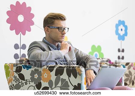 Mid-adult businessman using laptop on armchair in creative office View Large Photo Image Stock Image - Mid-adult businessman using laptop on armchair in creative office. Fotosearch