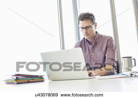 Stock Photo - Mid-adult businessman working laptop at desk in creative office. Fotosearch