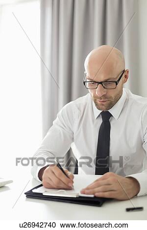 Stock Image - Mid adult businessman writing on clipboard in home office. Fotosearch