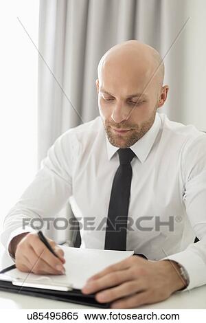 Stock Photography - Mid adult businessman writing on clipboard in home office. Fotosearch