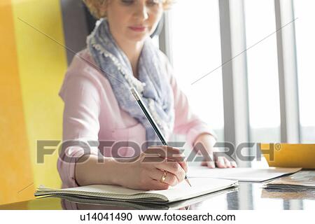 Stock Image - Mid adult businesswoman writing on notepad at desk in office. Fotosearch