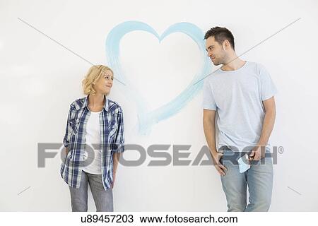 Stock Image - Mid-adult couple looking at each other with painted heart on wall. Fotosearch
