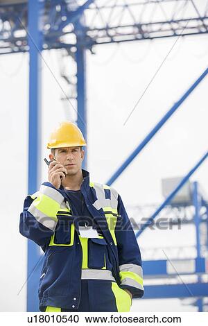 Stock Image - Mid adult male worker using walkie-talkie in shipping yard. Fotosearch