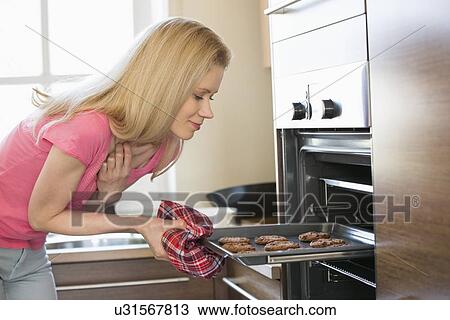 Stock Image - Mid adult woman removing baking tray from oven in kitchen. Fotosearch
