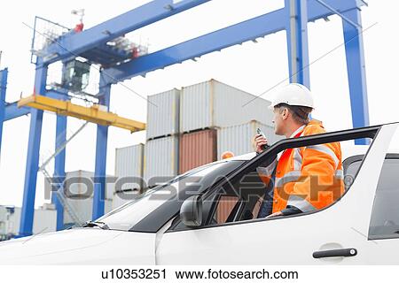 Stock Image - Middle-aged man using walkie-talkie while standing at car door in shipping yard. Fotosearch