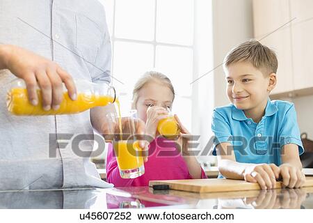 Stock Image - Midsection of father serving orange juice for children in kitchen. Fotosearch