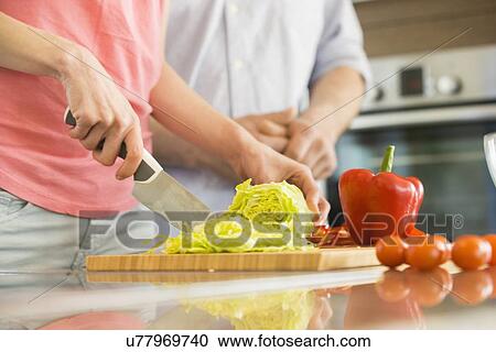 Stock Image - Midsection of woman chopping vegetables in kitchen with man standing in background. Fotosearch