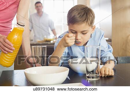 Stock Photo - Midsection of woman with juice bottle standing by son having breakfast with man in background. Fotosearch