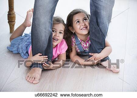 Stock Image - Playful girls holding father's legs on hardwood floor. Fotosearch