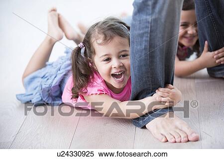 Stock Photography - Playful girls holding father's legs on hardwood floor. Fotosearch
