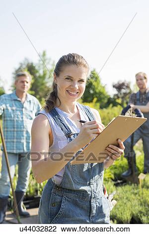 Portrait of beautiful supervisor holding clipboard with gardeners standing in background at plant nursery View Large Photo Image Stock Image - Portrait of beautiful supervisor holding clipboard with gardeners standing in background at plant nursery. Fotosearch