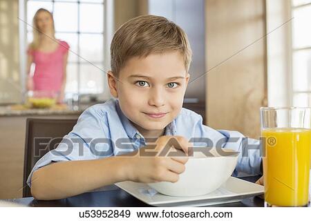 Portrait of boy having breakfast at table with mother standing in background View Large Photo Image Stock Photo - Portrait of boy having breakfast at table with mother standing in background. Fotosearch