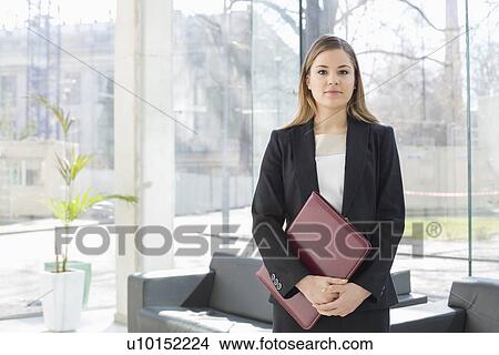 Portrait of businesswoman holding file while standing at office lobby View Large Photo Image Picture - Portrait of businesswoman holding file while standing at office lobby. Fotosearch