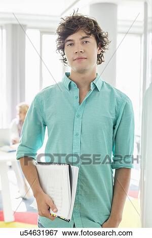 Stock Photo - Portrait of confident businessman holding files at creative office. Fotosearch