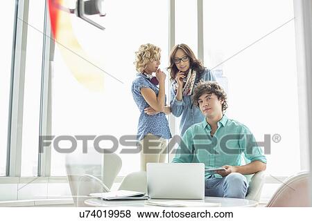 Portrait of confident businessman sitting at table with female colleagues conversing in background View Large Photo Image Stock Photo - Portrait of confident businessman sitting at table with female colleagues conversing in background. Fotosearch