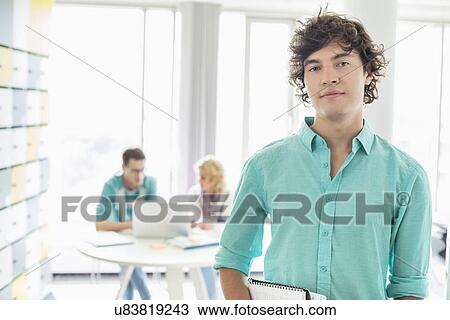 Stock Image - Portrait of confident businessman with colleagues working in background at creative office. Fotosearch