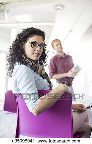 Portrait of confident businesswoman sitting on chair with colleague standing in background at creative office View Large Photo Image Stock Image - Portrait of confident businesswoman sitting on chair with colleague standing in background at creative office. Fotosearch
