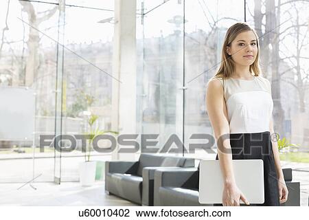 Portrait of confident businesswoman holding laptop while standing at office lobby View Large Photo Image Stock Image - Portrait of confident businesswoman holding laptop while standing at office lobby. Fotosearch