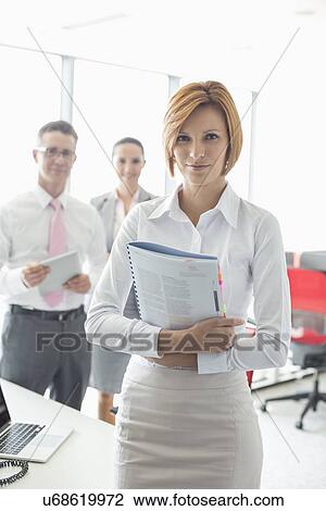 Portrait of confident businesswoman holding book with colleagues in background at office View Large Photo Image Stock Image - Portrait of confident businesswoman holding book with colleagues in background at office. Fotosearch