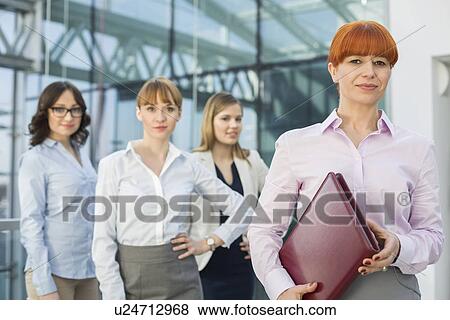 Stock Photo - Portrait of confident businesswoman holding folder with female colleagues standing in background. Fotosearch