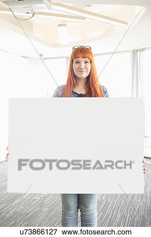 Stock Photo - Portrait of confident businesswoman holding blank placard in creative office. Fotosearch
