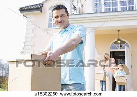 Stock Image - Portrait of confident man carrying cardboard box while moving house with family in background. Fotosearch
