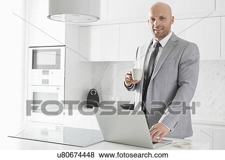 Portrait of confident mid adult businessman having coffee while using laptop in kitchen View Large Photo Image Stock Photo - Portrait of confident mid adult businessman having coffee while using laptop in kitchen. Fotosearch