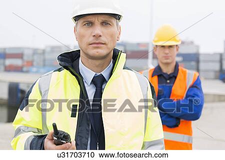 Portrait of confident worker standing with coworker in background at shipping yard View Large Photo Image Stock Image - Portrait of confident worker standing with coworker in background at shipping yard. Fotosearch