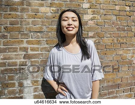 Picture - Portrait of confident young woman standing with hand on hip against brick wall. Fotosearch