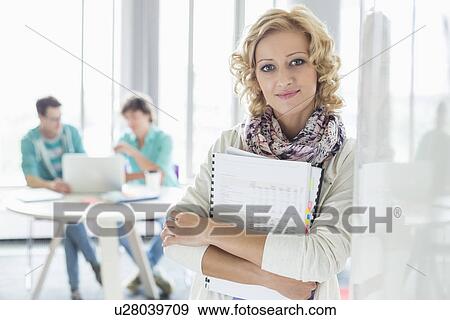Portrait of creative businesswoman holding files with colleagues working in background at office View Large Photo Image Stock Photo - Portrait of creative businesswoman holding files with colleagues working in background at office. Fotosearch