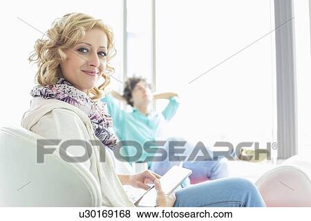 Stock Photo - Portrait of creative businesswoman holding digital tablet at lobby with colleague relaxing in background. Fotosearch
