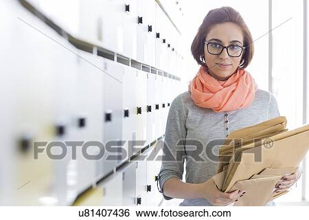 Stock Photograph - Portrait of creative businesswoman holding envelopes in locker room at office. Fotosearch