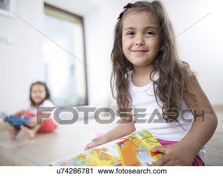 Portrait of cute girl holding book with sister in background at home View Large Photo Image Stock Image - Portrait of cute girl holding book with sister in background at home. Fotosearch