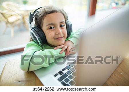 Stock Image - Portrait of cute girl listening to music on headphones with laptop at table. Fotosearch