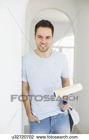 Stock Image - Portrait of handsome man holding paint roller in new house. Fotosearch