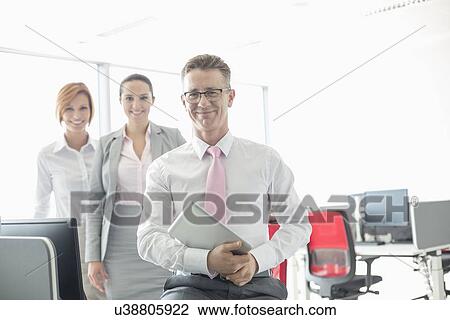 Portrait of happy businessman holding tablet PC with female colleagues in background at office View Large Photo Image Stock Image - Portrait of happy businessman holding tablet PC with female colleagues in background at office. Fotosearch