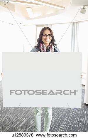 Portrait of happy businesswoman holding blank sign in creative office View Large Photo Image Stock Photo - Portrait of happy businesswoman holding blank sign in creative office. Fotosearch