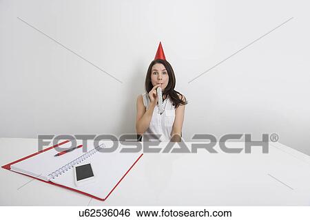 Portrait of happy businesswoman in party hat blowing horn at desk in office View Large Photo Image Stock Photograph - Portrait of happy businesswoman in party hat blowing horn at desk in office. Fotosearch