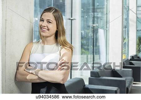 Stock Photograph - Portrait of happy businesswoman standing arms crossed at office lobby. Fotosearch