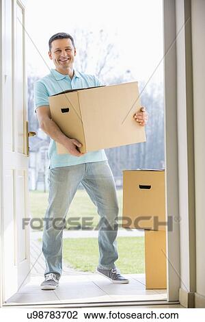 Stock Image - Portrait of happy man carrying cardboard box while entering new home. Fotosearch